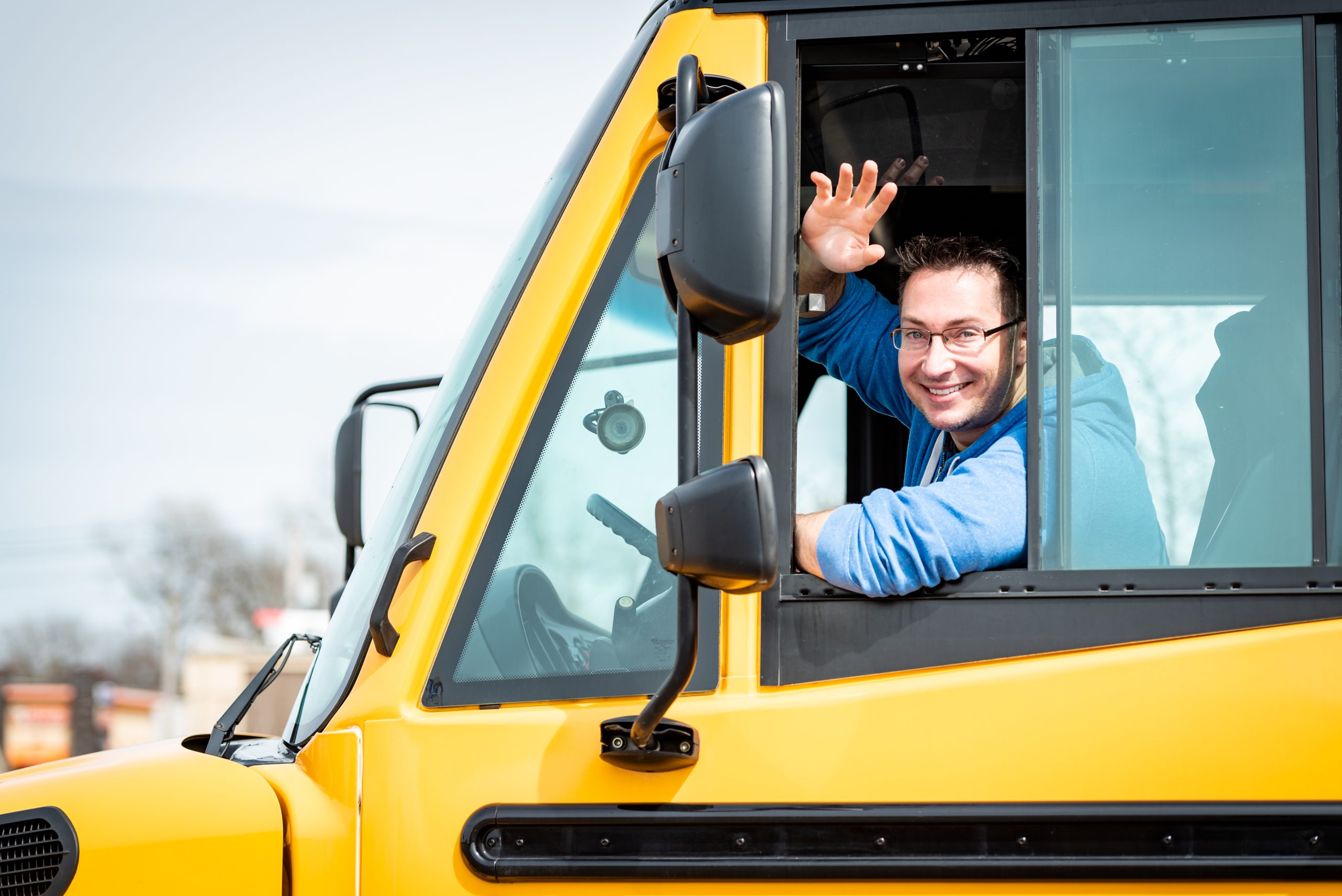School bus driver smiling and waving from the driver’s seat.